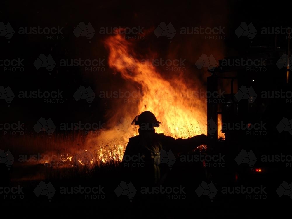 Firefighter fighting a grass fire at night silhouetted against the fire - Australian Stock Image