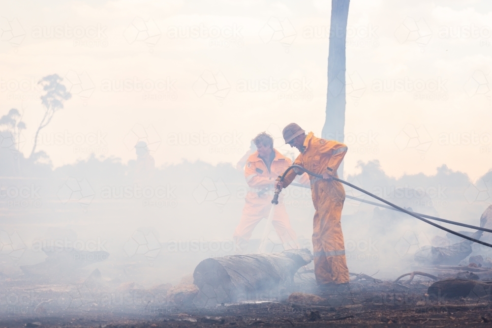 firefighters using hoses to spray water on burnt logs after a fire - Australian Stock Image