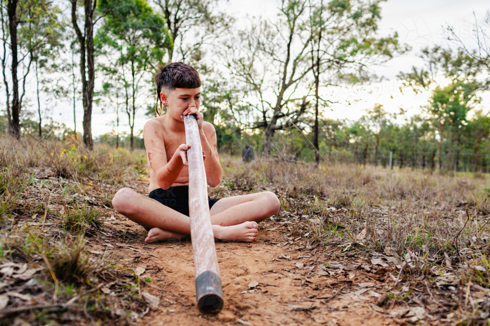 First Nations Australian boy playing didgeridoo in Aussie bushland painted with ochre - Australian Stock Image