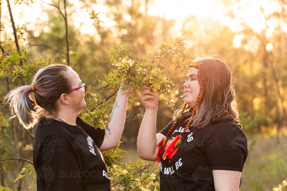First Nations Australian mother and teen daughter with wattle flowers in bushland - Australian Stock Image