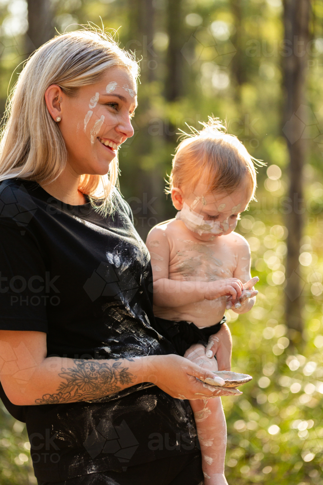 First Nations Australian woman with baby boy applying cultural ochre body paint mixed in shell - Australian Stock Image