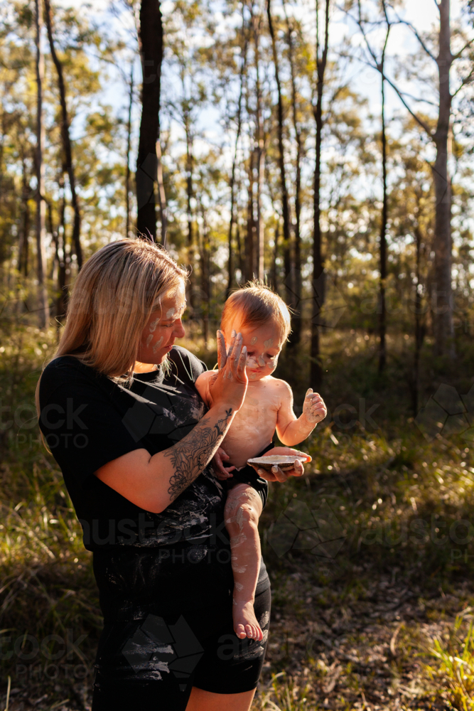 First Nations Australian woman with baby boy applying cultural ochre body paint mixed in shell - Australian Stock Image