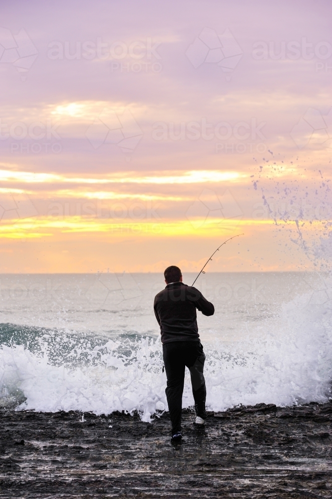 Fisherman on the rocks with waves crashing up at sunrise - Australian Stock Image