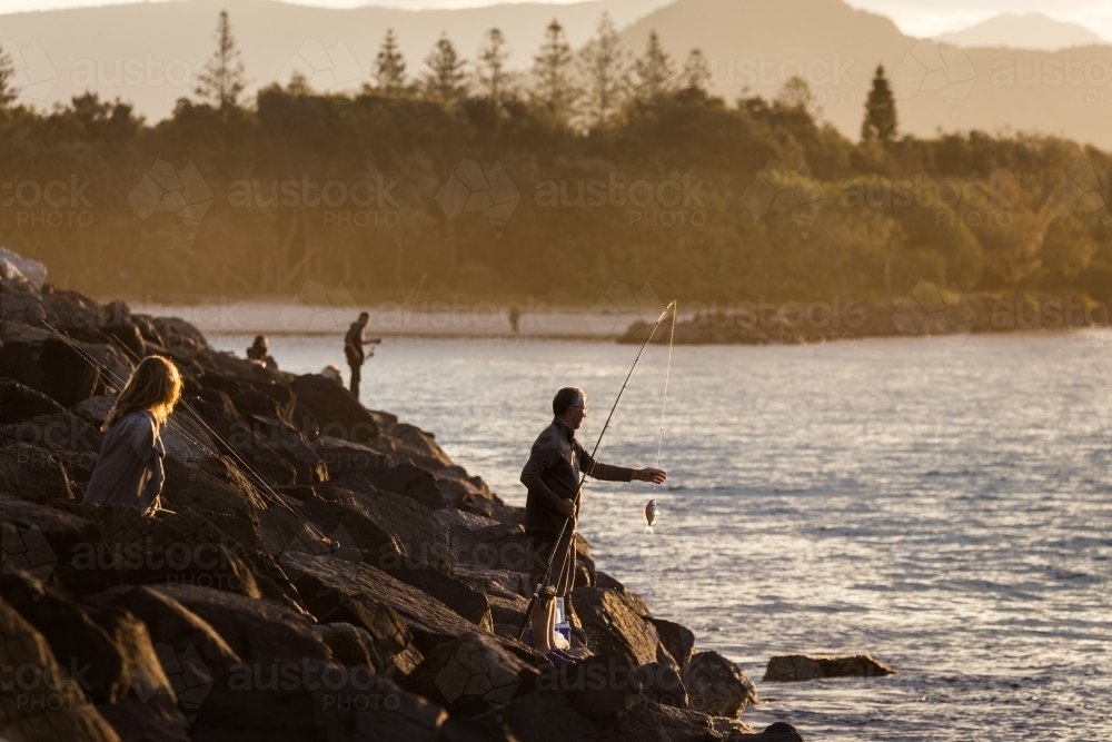 Fisherman standing on rocks beside a river at sunset - Australian Stock Image