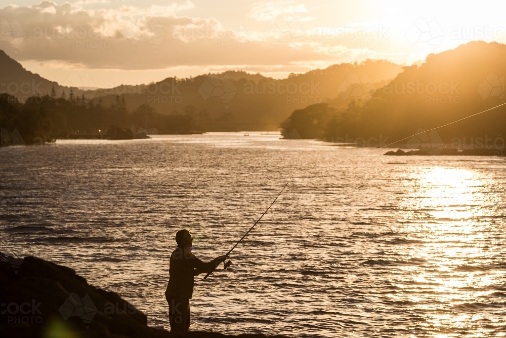 Fisherman standing on rocks beside a river at sunset - Australian Stock Image