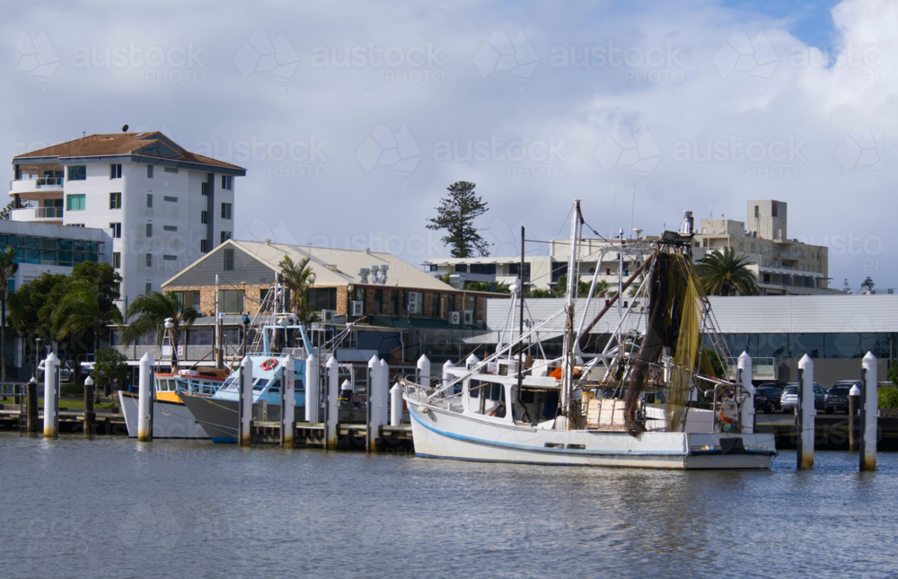 Fishing boats moored in river with waterfront buildings on shoreline - Australian Stock Image