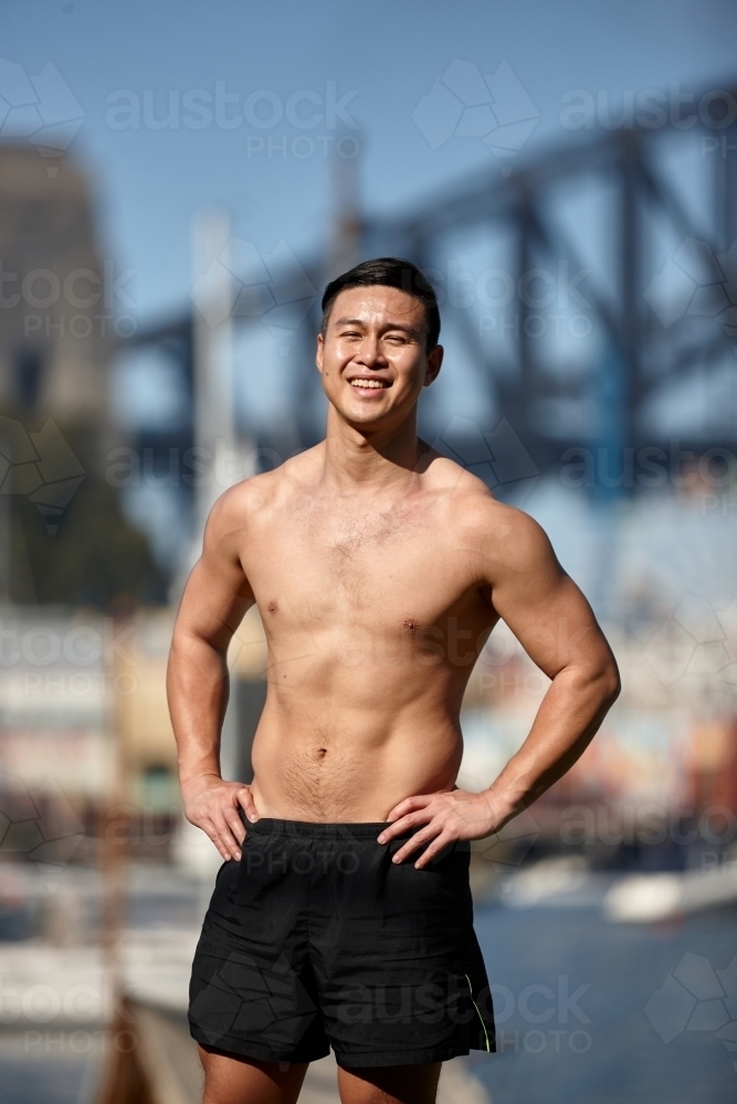 Fit Asian man exercising and smiling in front of harbour bridge - Australian Stock Image