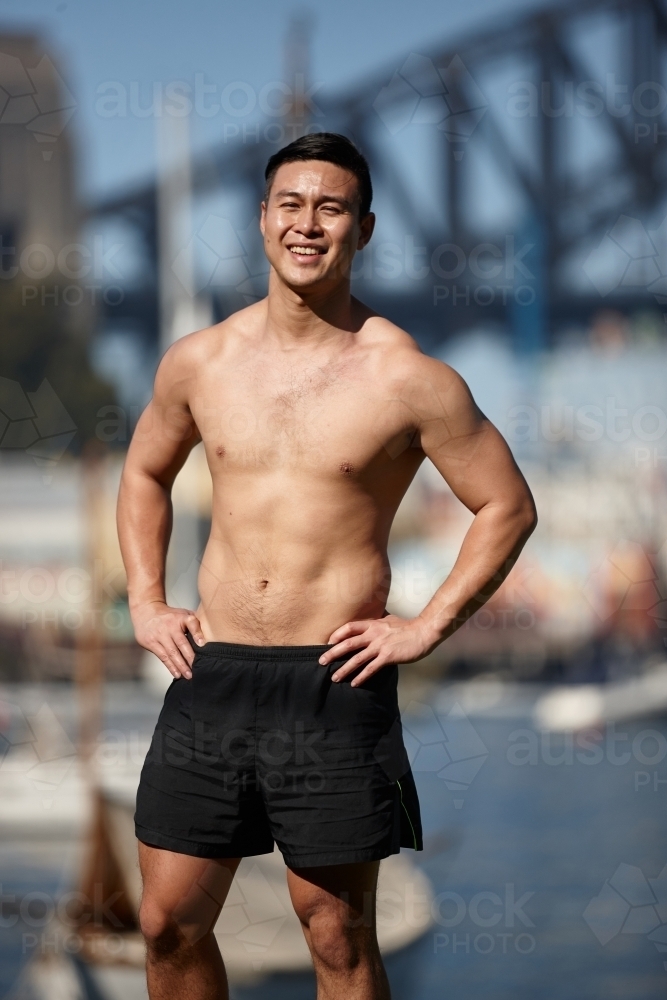 Fit Asian man smiling with hands on hips in front of harbour bridge - Australian Stock Image