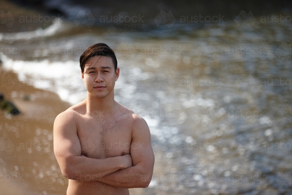 Fit Asian man with arms folded at beach - Australian Stock Image