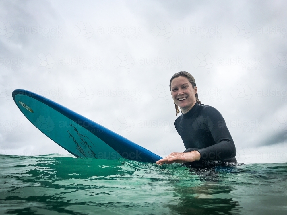 Fit, healthy natural woman sitting on longboard surfboard smiling at camera on overcast day - Australian Stock Image
