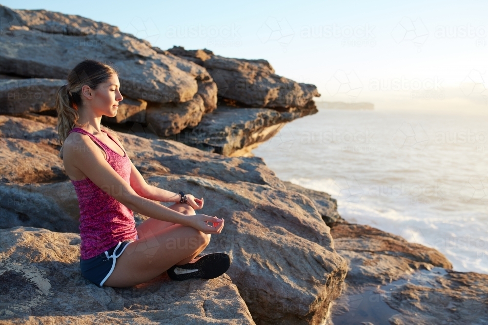 Fitness woman meditating at coastal headland on sunrise - Australian Stock Image