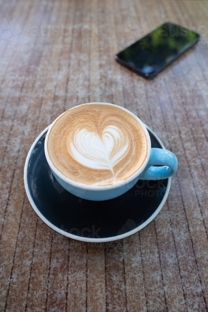 flat white coffee in a wide blue coffee cup with black saucer, with a phone in the background - Australian Stock Image