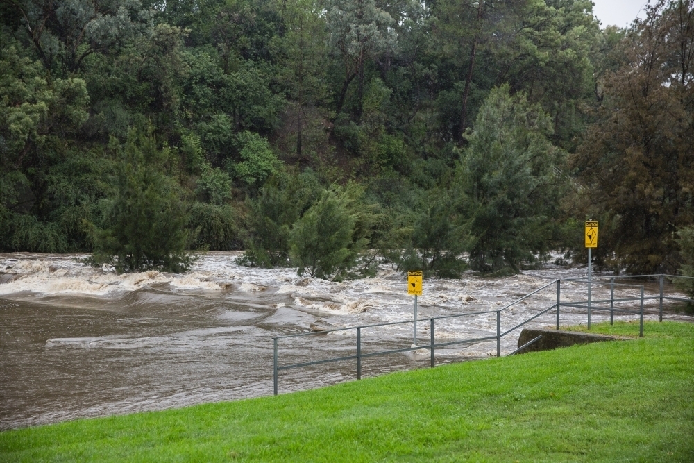 Flooded river water rushing downstream with trees and signs - Australian Stock Image