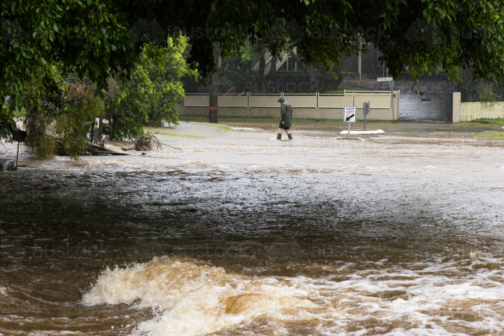 Floodwaters rushing across a road with a distant person walking through water in the background - Australian Stock Image