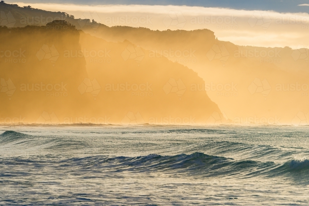 Fog rising above waves in front of sea cliffs glowing orange in dawn sunshine - Australian Stock Image