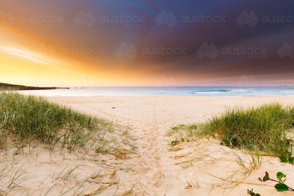 Footprints in the sand on a beach walkway lead towards a beautiful, dramatic sunrise over the sea - Australian Stock Image