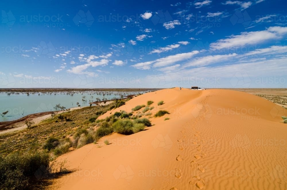 Footprints on a sand dune leading to a man and a vehicle with lake below and cloud formations - Australian Stock Image
