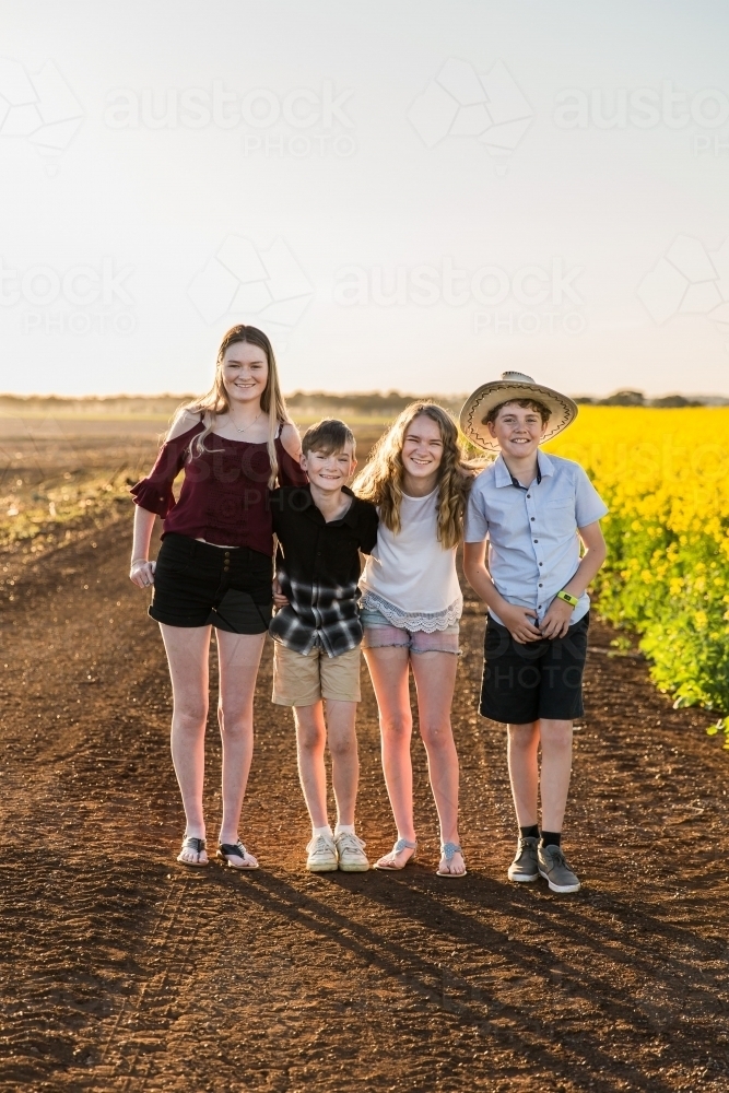 Four children from the same family standing together happy on farm on dirt road next to canola - Australian Stock Image