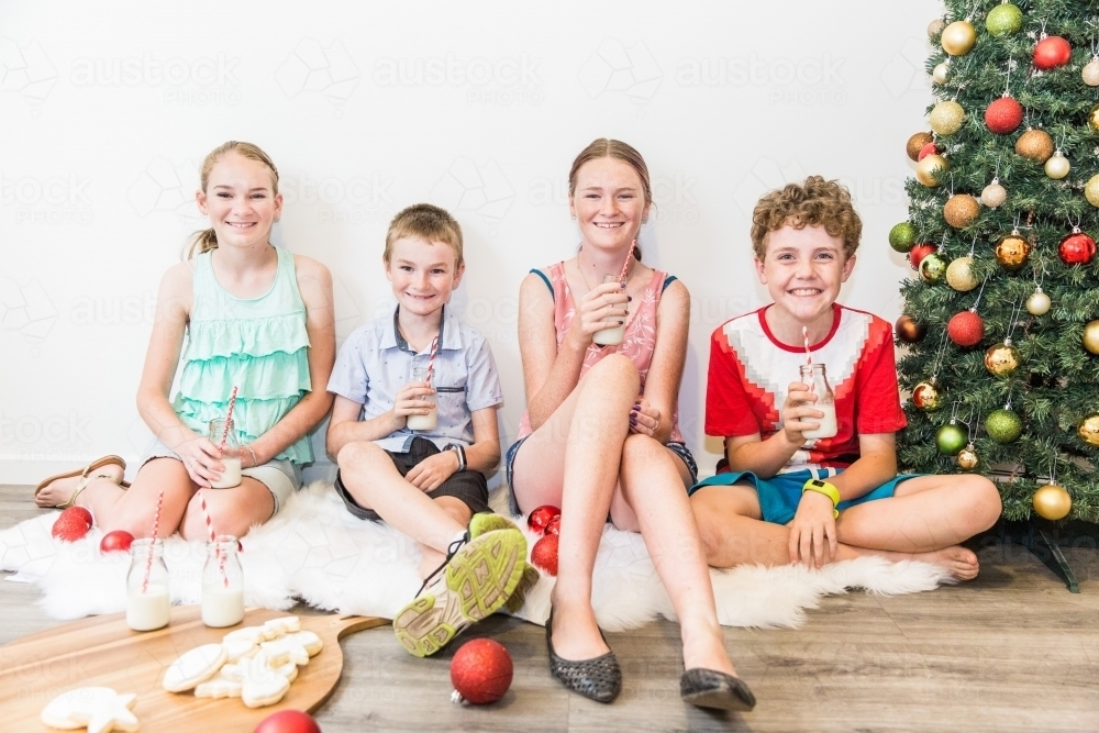 Four children sitting on rug next to Christmas tree having milk and cookies - Australian Stock Image