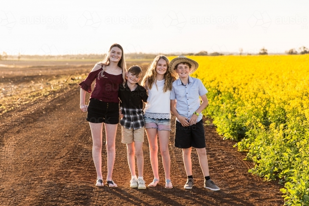 Four children standing close together happy smiling on farm near canola field - Australian Stock Image