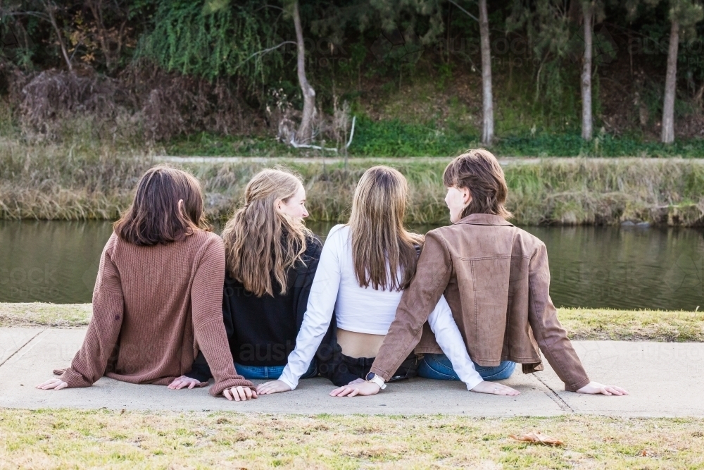 Four friends sitting on path near river leaning back on hands - Australian Stock Image