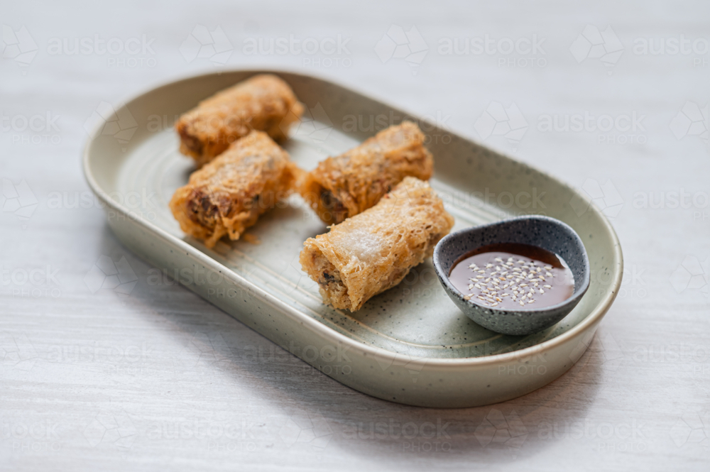 Four golden crispy spring rolls on plate alongside a small bowl of sauce - Australian Stock Image