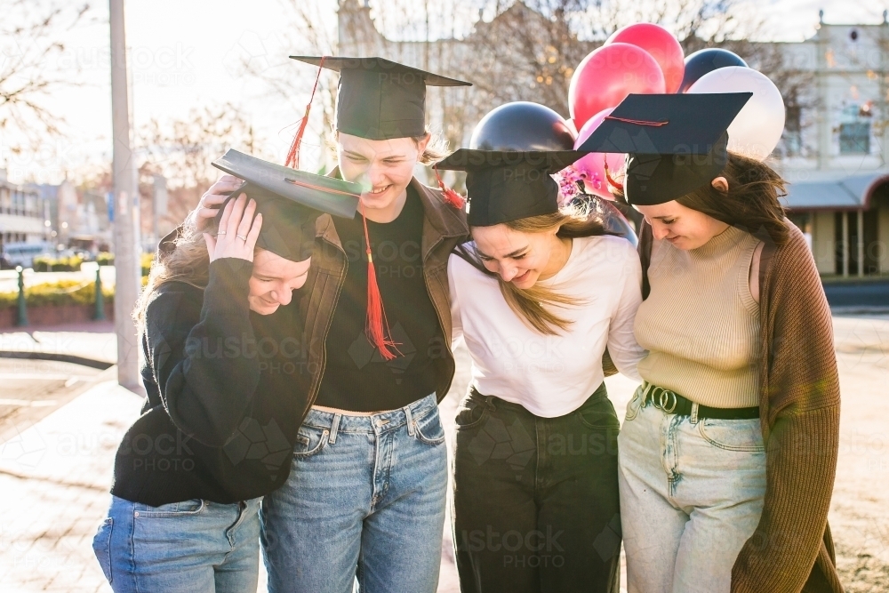 Four teenage girls standing together wearing graduation caps laughing with balloons - Australian Stock Image