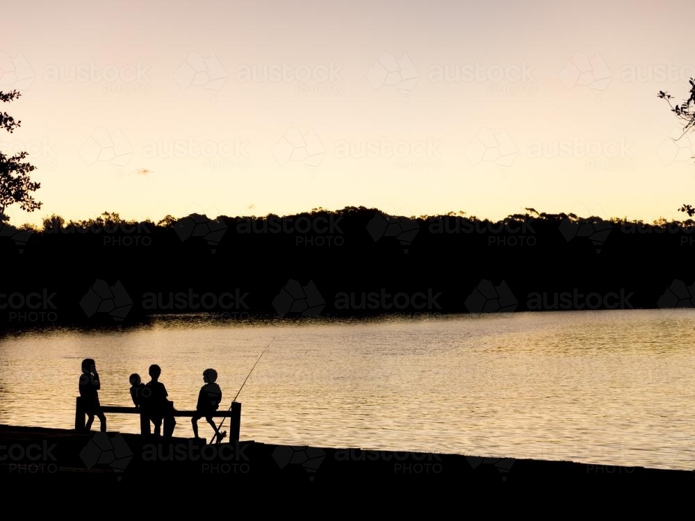 Four young children silhouetted against water with fishing rod - Australian Stock Image