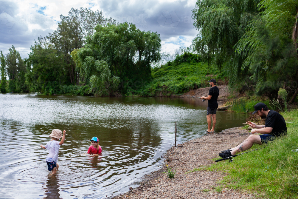 Friends together by river with kids playing in water and men fishing on summer afternoon - Australian Stock Image