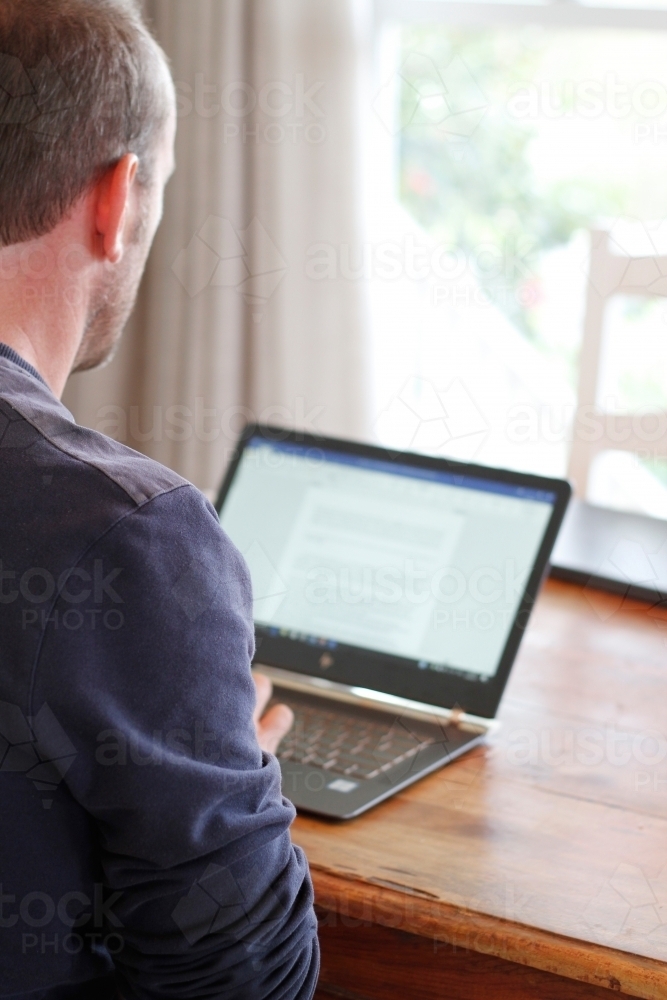 From behind view of man working on laptop computer at table - Australian Stock Image