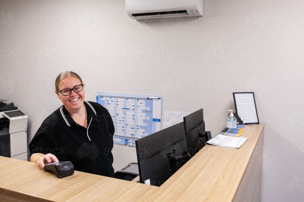 front desk lady smiling while holding out payment terminal at mechanics office - Australian Stock Image