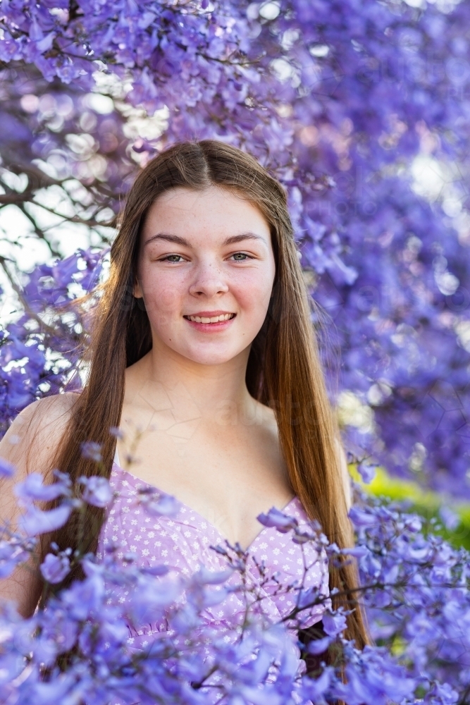 Front on portrait of a happy smiling teen girl with long brown hair among purple jacaranda flowers - Australian Stock Image
