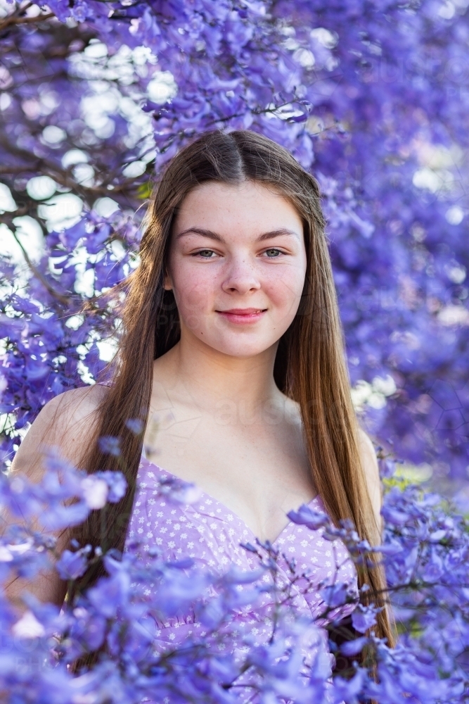 Front on portrait of a happy smiling teen girl with long brown hair among purple jacaranda flowers - Australian Stock Image