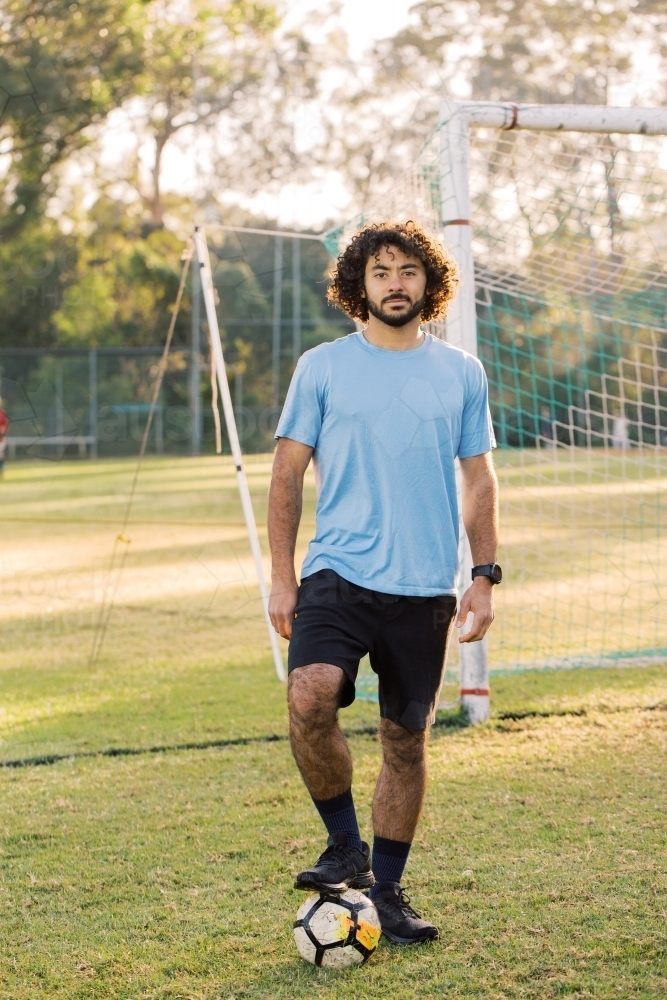 full body shot of a man with curly hair stepping over a soccer ball with one foot on a field - Australian Stock Image