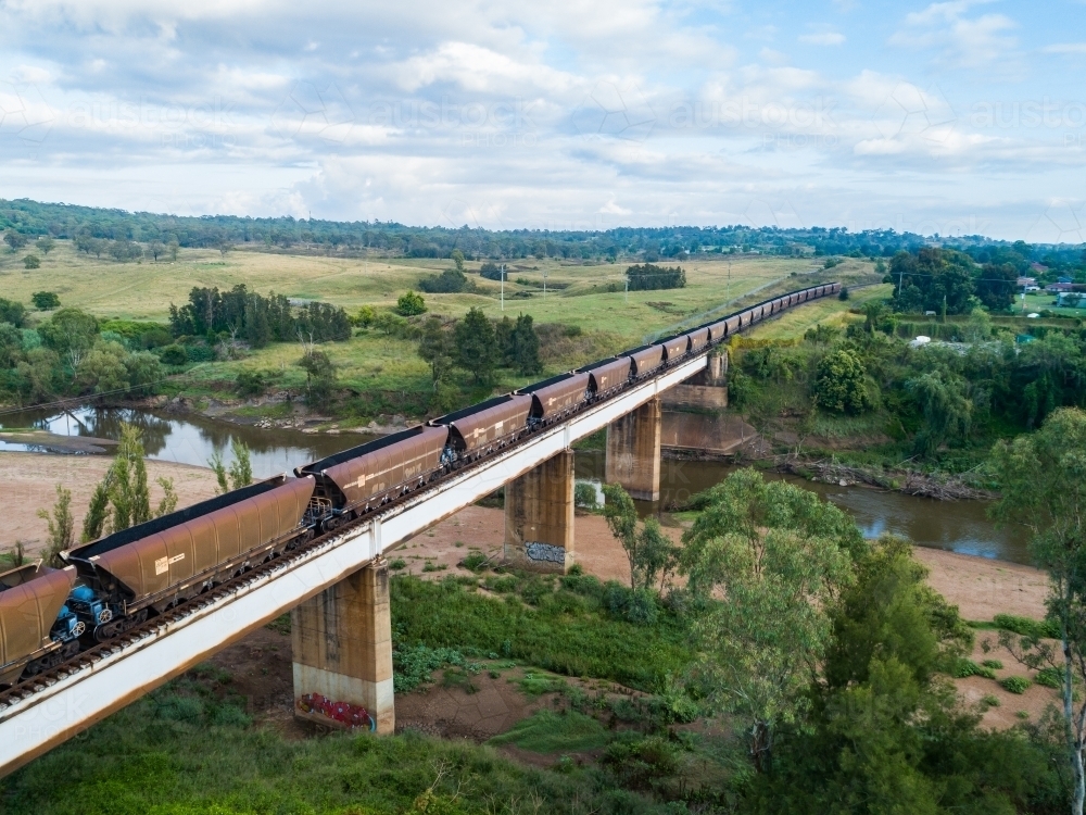 Full coal train traveling over railway bridge on overcast morning - Australian Stock Image