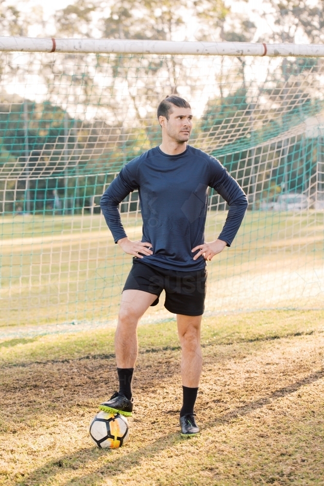 Full length man standing on the field stepping on a soccer ball near the soccer net - Australian Stock Image