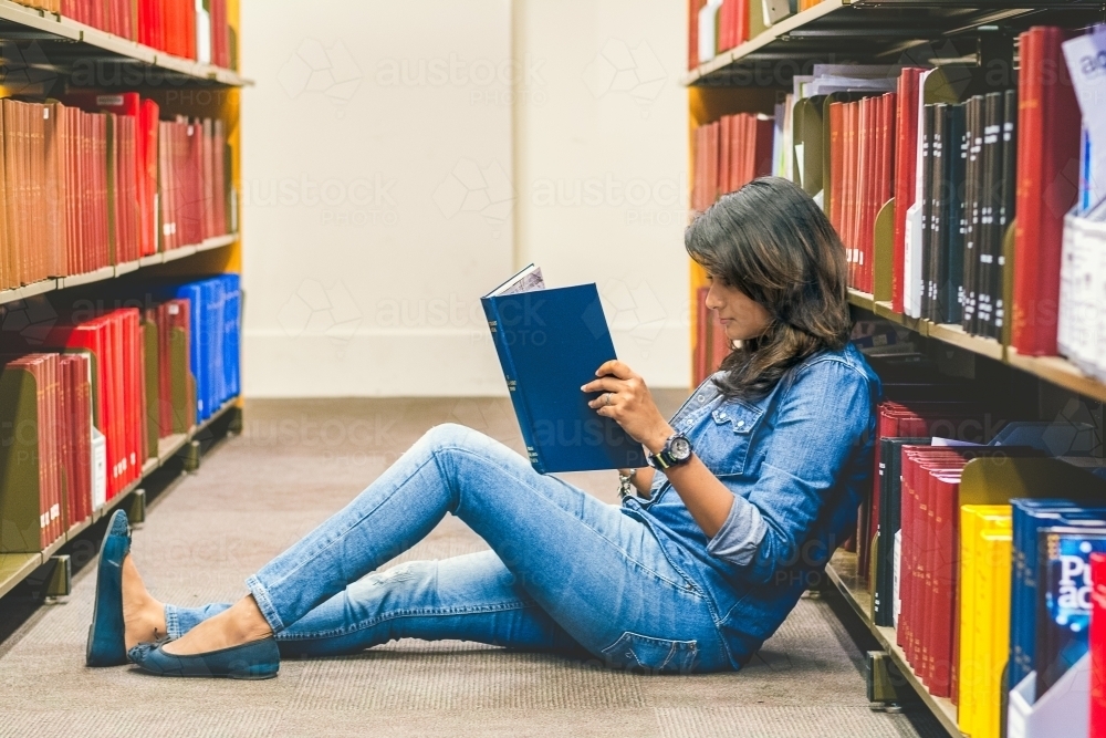 Full Length Of Woman Reading Book Amidst Shelves At Library - Australian Stock Image