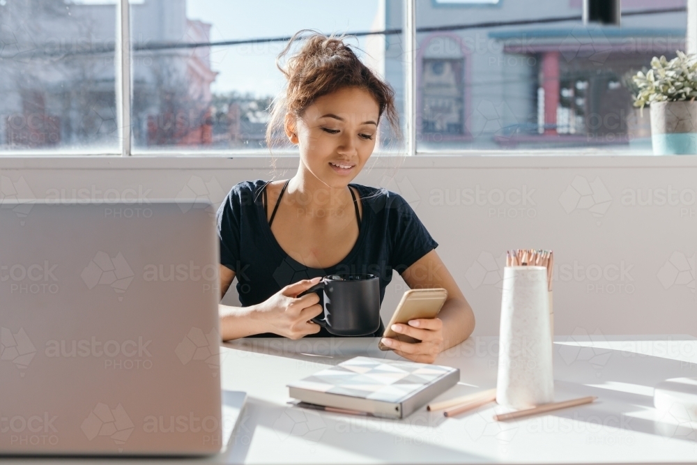 Gen Y female engaging on social media on her phone while at her desk - Australian Stock Image