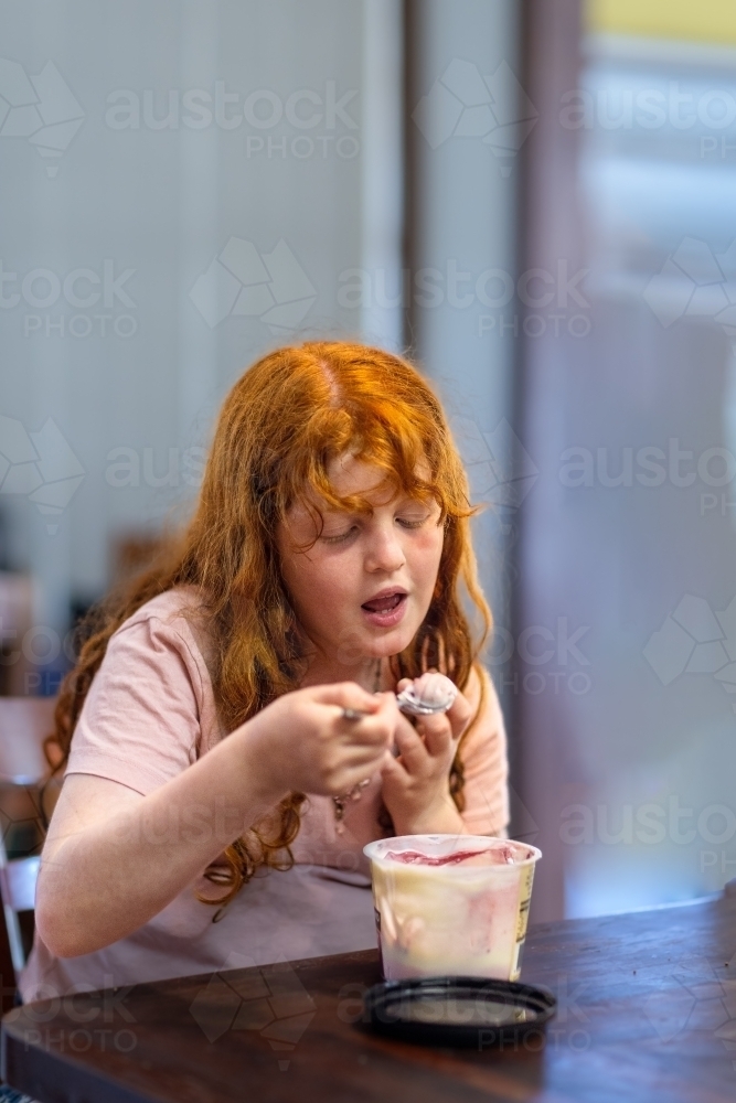 ginger-haired girl eating flavoured yoghurt on a spoon from plastic tub - Australian Stock Image