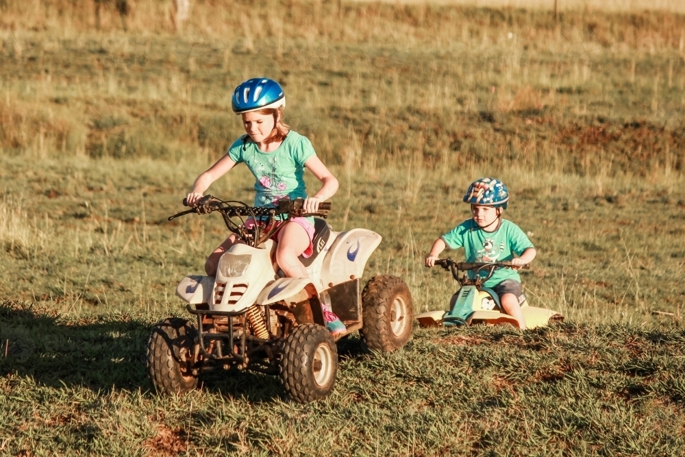 Girl and boy riding quad bikes over contour bank on farm property - Australian Stock Image