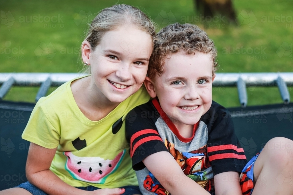 Girl and boy sitting close together on trampoline smiling - Australian Stock Image