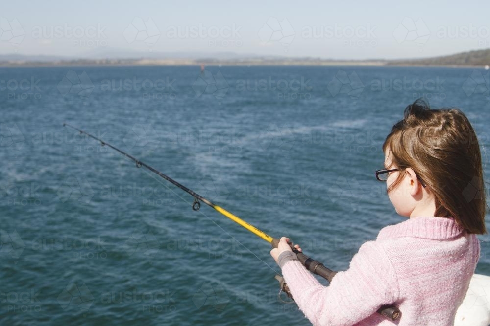 Girl fishing off wharf on windy sunlit day - Australian Stock Image