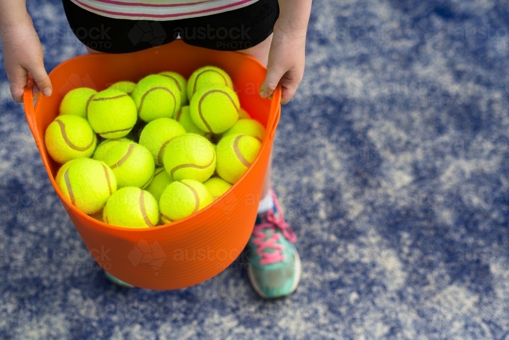 Girl holding an orange basket of tennis balls on a blue tennis court - Australian Stock Image