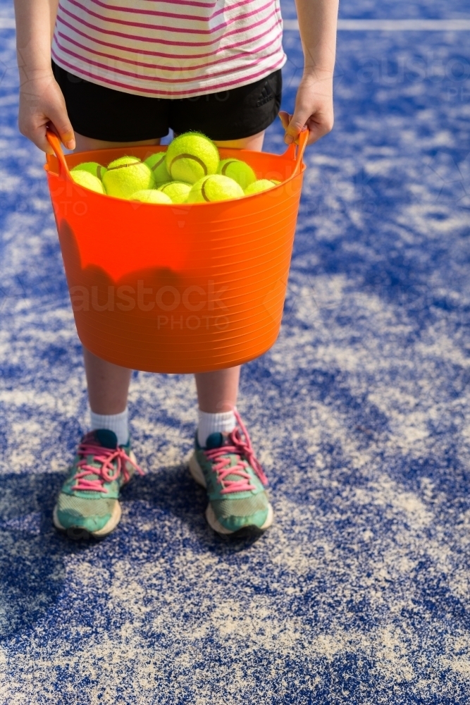 Girl holding an orange basket of tennis balls on a blue tennis court - Australian Stock Image