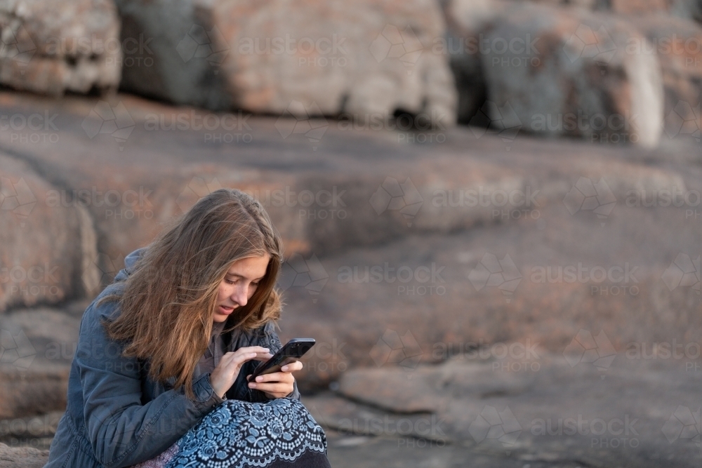 Girl huddled in jacket outdoors looking at smartphone - Australian Stock Image
