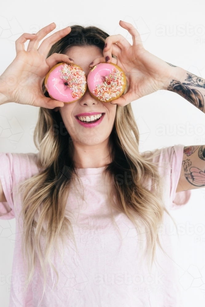 Girl in a pink t shirt holding pink donuts up to her eyes - Australian Stock Image
