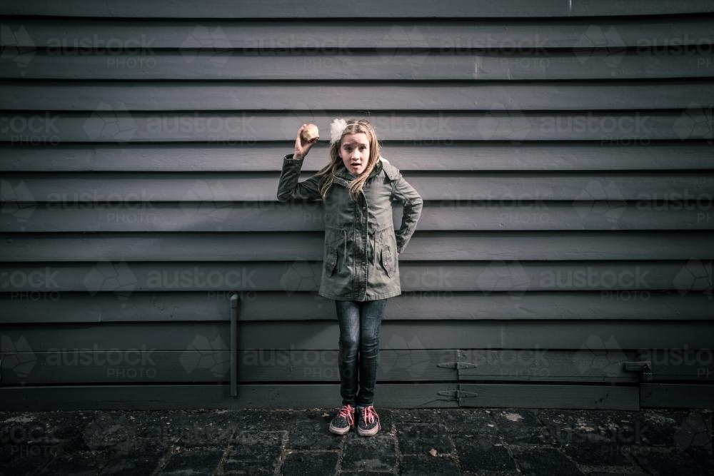 Girl in grey standing in front of grey weatherboards in the city holding an apple - Australian Stock Image