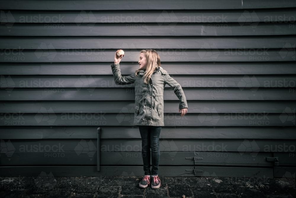 Girl in grey standing in front of grey weatherboards in the city holding an apple - Australian Stock Image