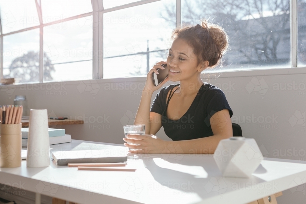 Girl in her 20s talking on a mobile phone at a desk in a light bright office - Australian Stock Image