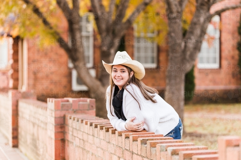 Girl leaning on brick wall near building smiling in hat and scarf - Australian Stock Image
